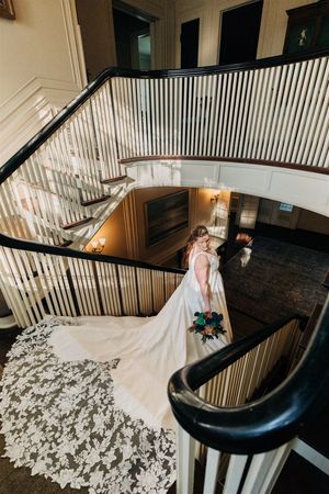 Elegant wedding gown with intricate lace train cascading down curved staircase with dark wooden banister.