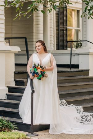 Beautiful bride in flowing white gown holds vibrant orange and green bouquet while standing on historic home's porch steps.