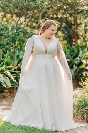 A white lace wedding gown with tulle details and floral embellishments is photographed outdoors in a garden setting.