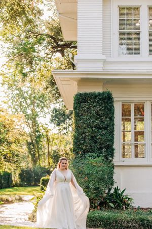 Elegant wedding gown flowing in breeze against backdrop of white colonial house with manicured landscaping.