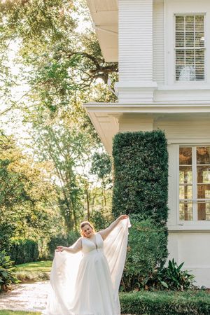 Elegant white wedding gown flowing in sunlight against classic white mansion with ivy-covered walls and lush gardens.