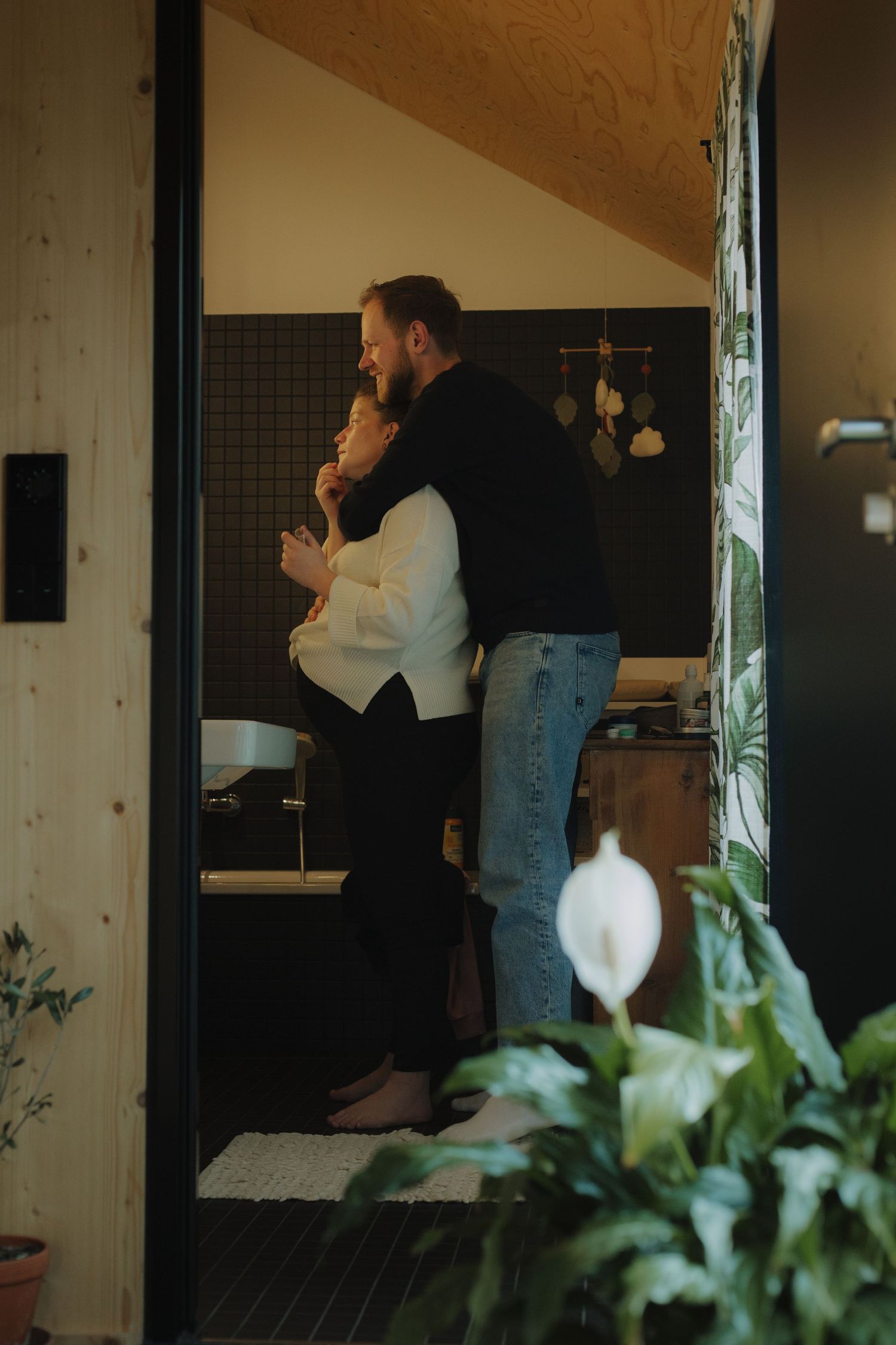 Pregnant Couple embracing in doorway of modern bath with indoor plants.