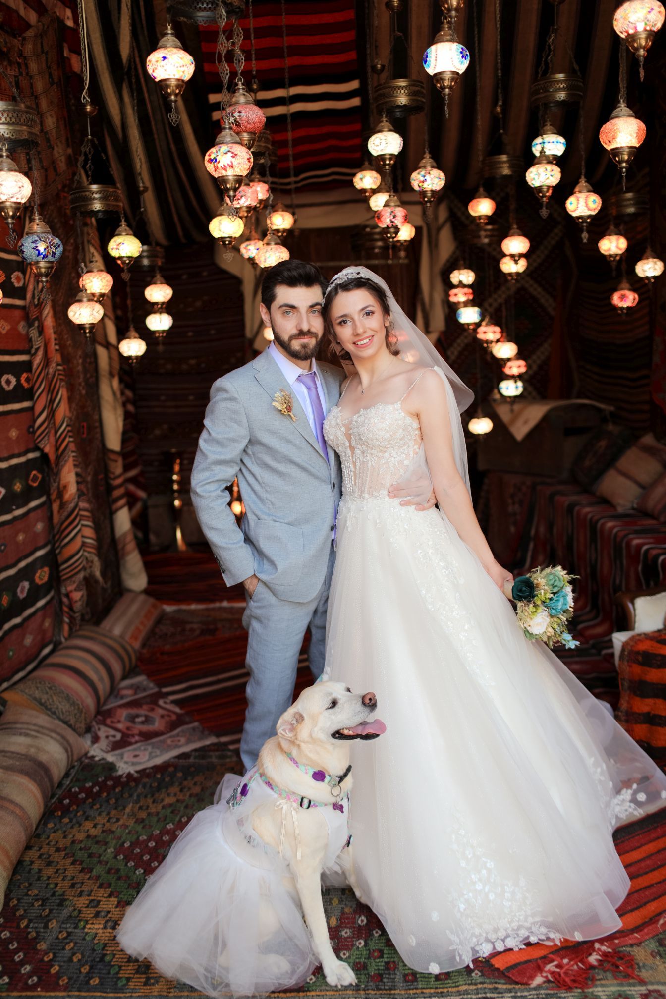 A couple poses in their wedding attire with their white dog under ornate Turkish lanterns in a richly decorated interior space.