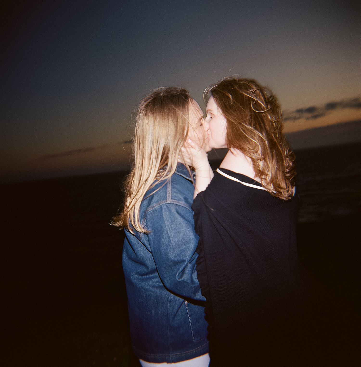 Two people in casual outfits share an intimate moment at sunset against a darkening sky.