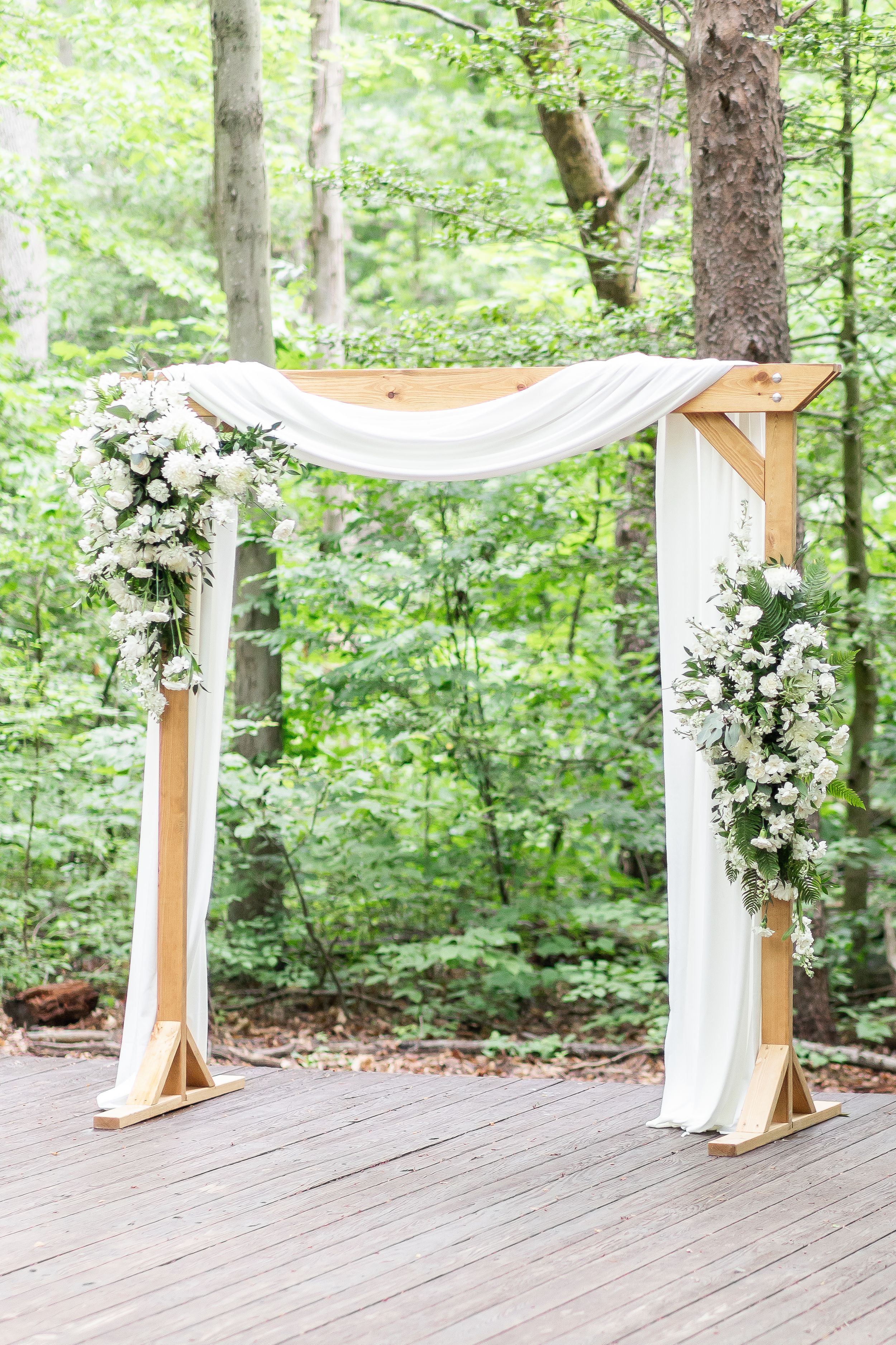 Elegant white wedding arch decorated with flowing fabric and white floral arrangements in a wooded outdoor setting.