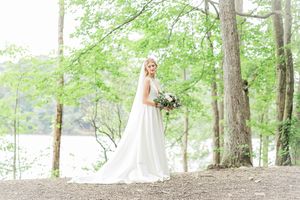 A person in a white wedding dress stands holding a bouquet in a sunlit forest setting beside a lake.