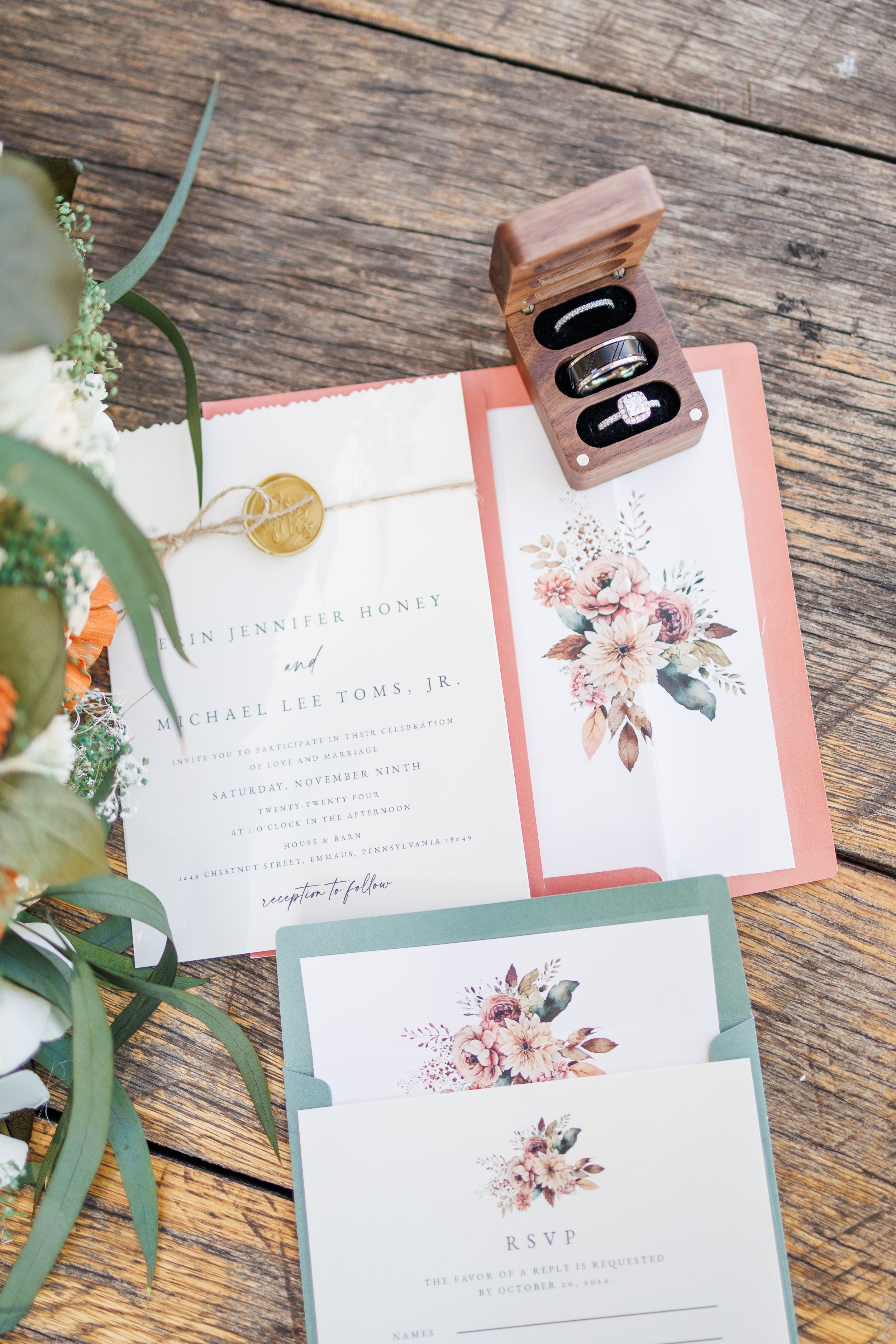 Wedding invitation suite with floral designs and gold details alongside rings in wooden box on rustic wood surface.