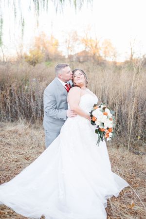 Wedding couple embraces in field of tall grass at sunset wearing gray suit and white gown with peach bouquet.