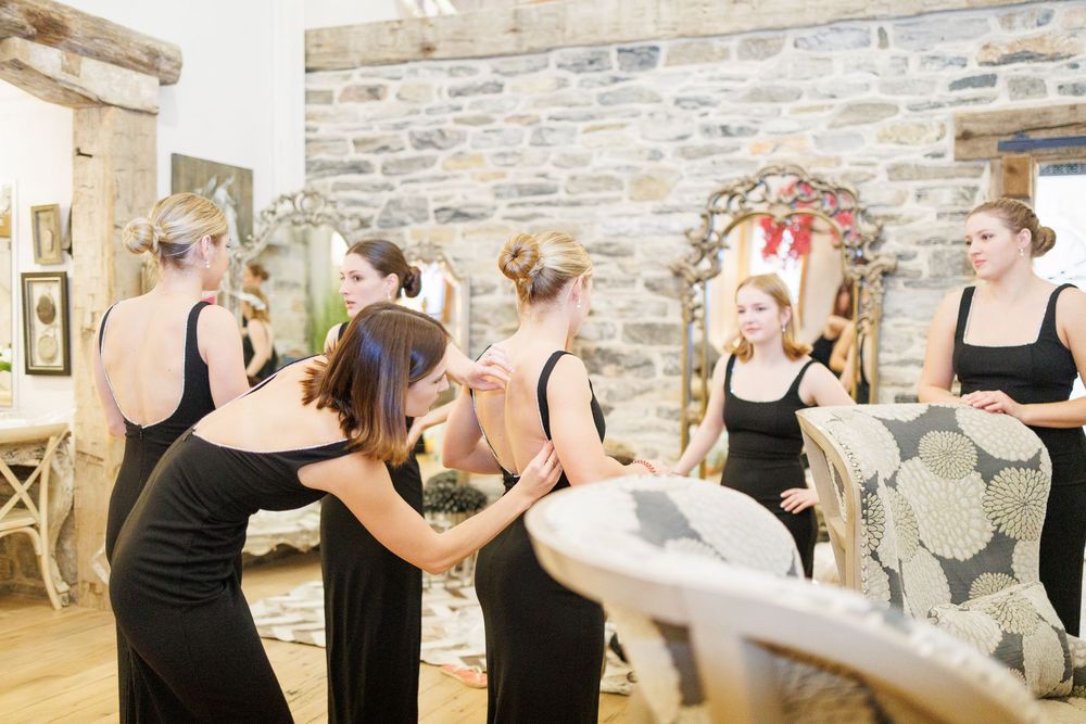 Bridesmaids in black dresses help each other prepare in a rustic stone-walled room before the wedding ceremony.
