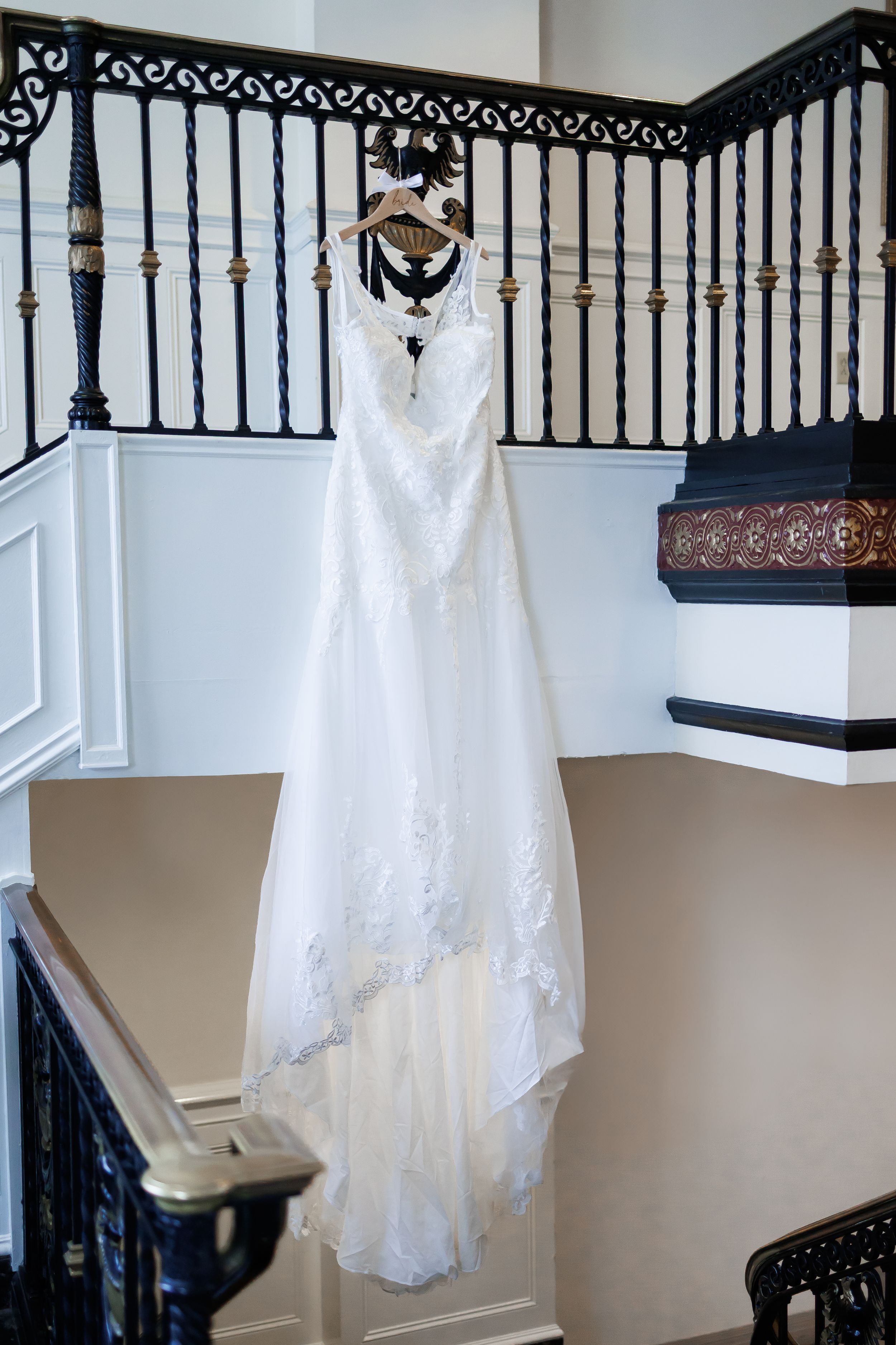 A white lace wedding dress hangs elegantly on display against an ornate black staircase railing.