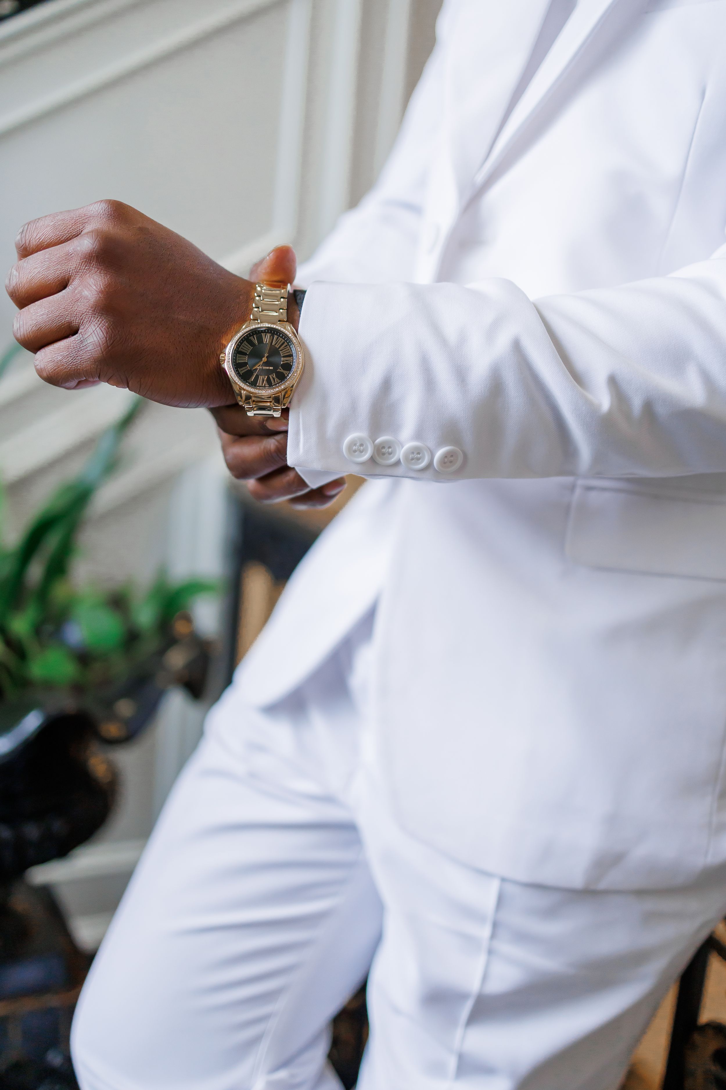 Person in crisp white suit adjusts elegant gold watch on wrist while standing indoors near plants.