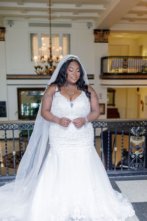 A bride in an elegant white lace mermaid wedding gown with a long veil poses in an ornate hotel lobby setting.