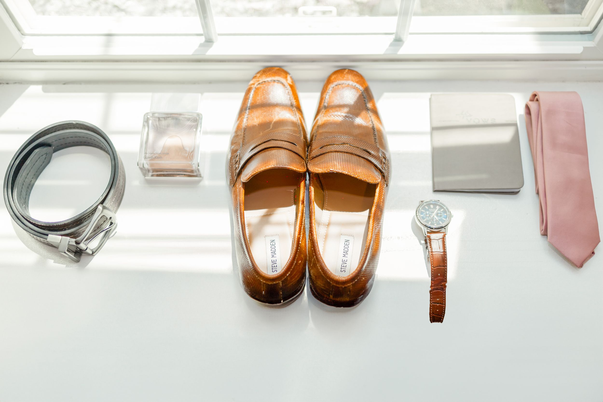 Brown leather dress shoes arranged with accessories including a gray belt, watch, pink tie and perfume bottle on white surface.