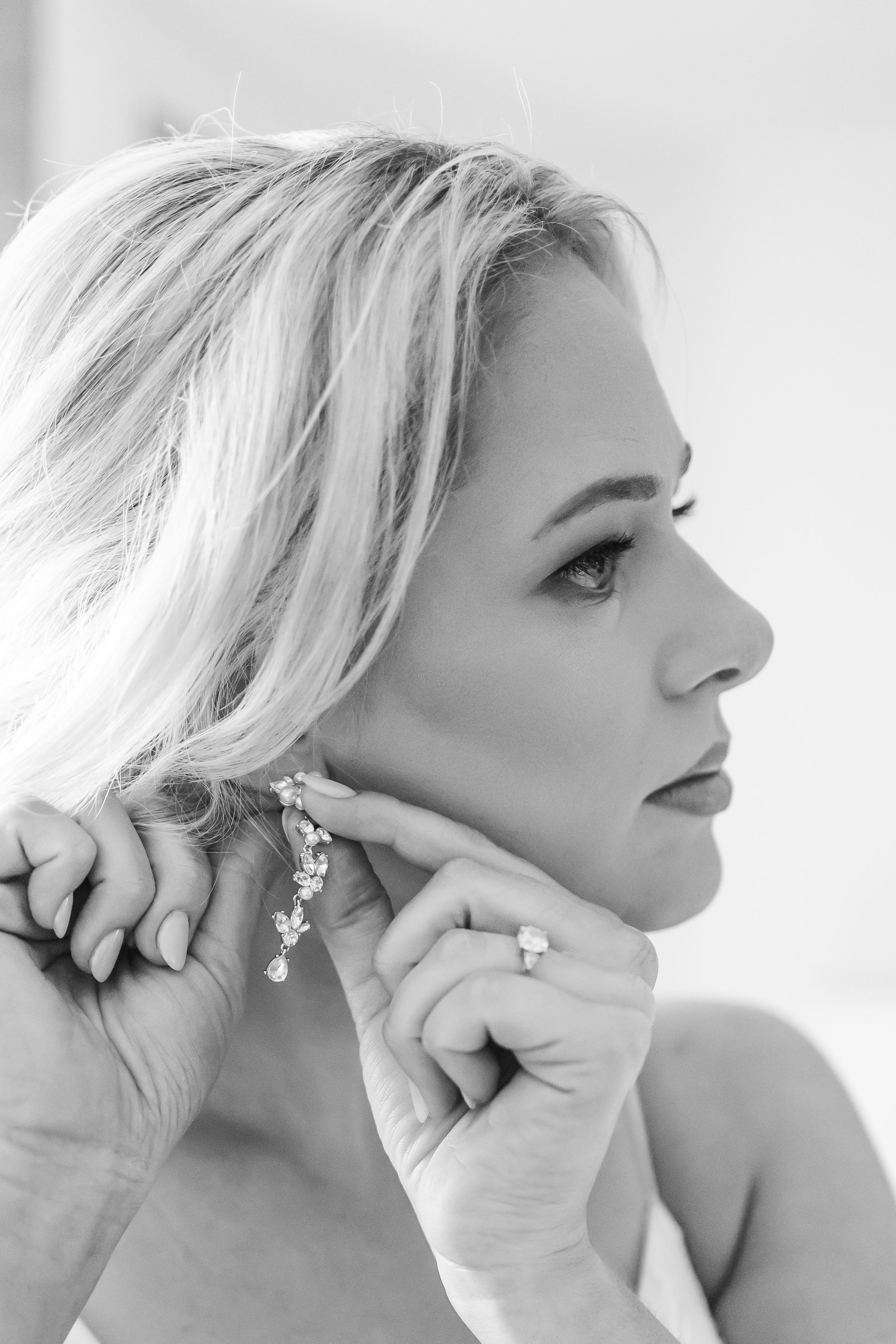 Black and white profile portrait of a woman with blonde hair wearing elegant drop earrings and rings in soft lighting.