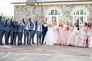Wedding party in classic blue suits and pink dresses poses together outside stone building with arched windows.