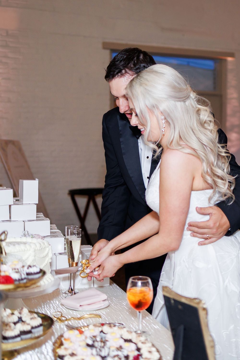 Newlywed couple cutting their wedding cake together at reception with drinks and dessert table in background.