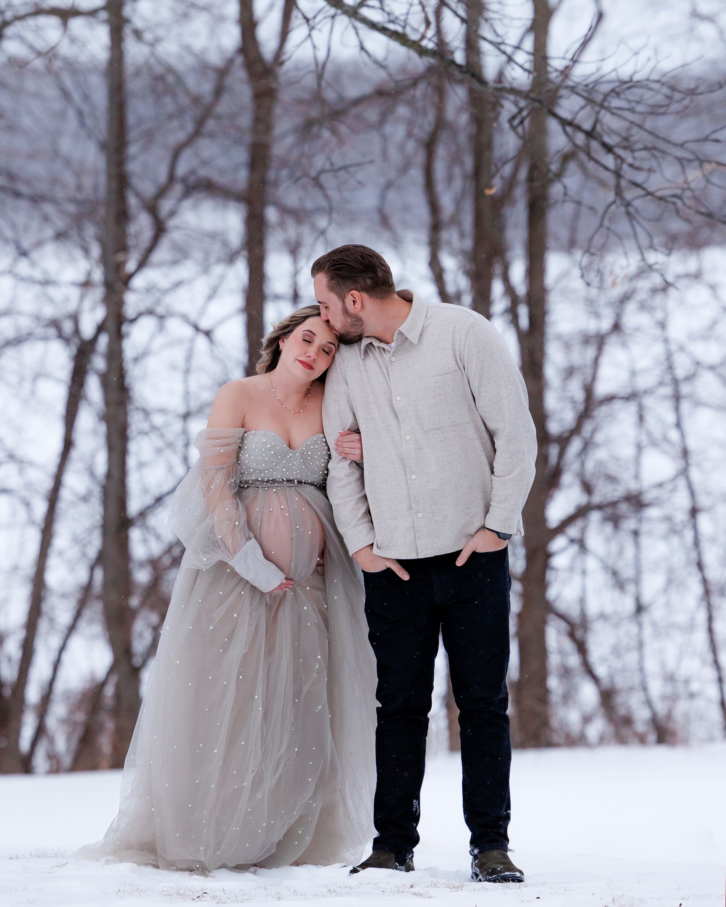 A couple shares an intimate moment in a snowy winter forest setting, with the woman wearing an elegant grey gown.