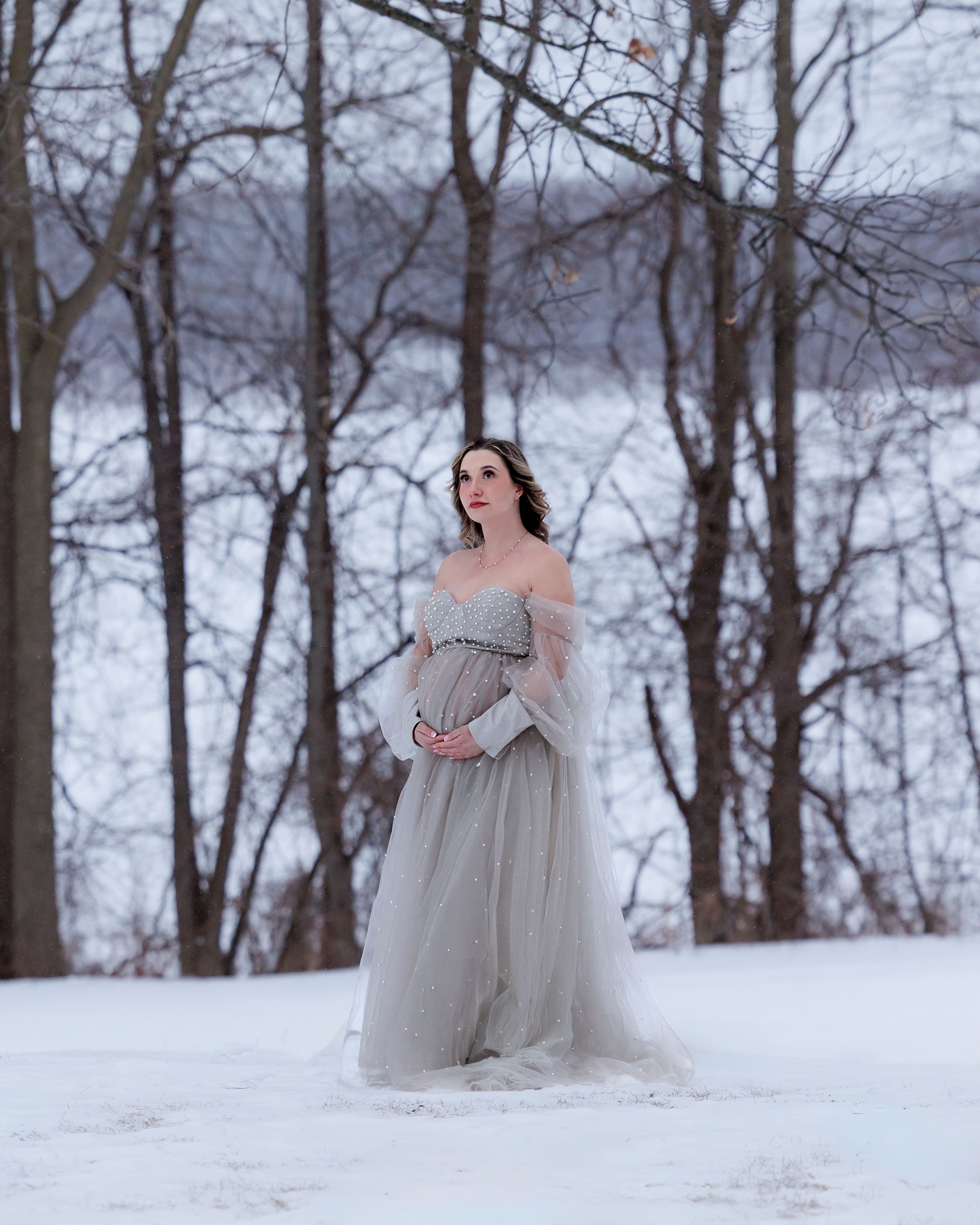 A woman in a flowing gray dress stands gracefully in a snowy winter forest setting.