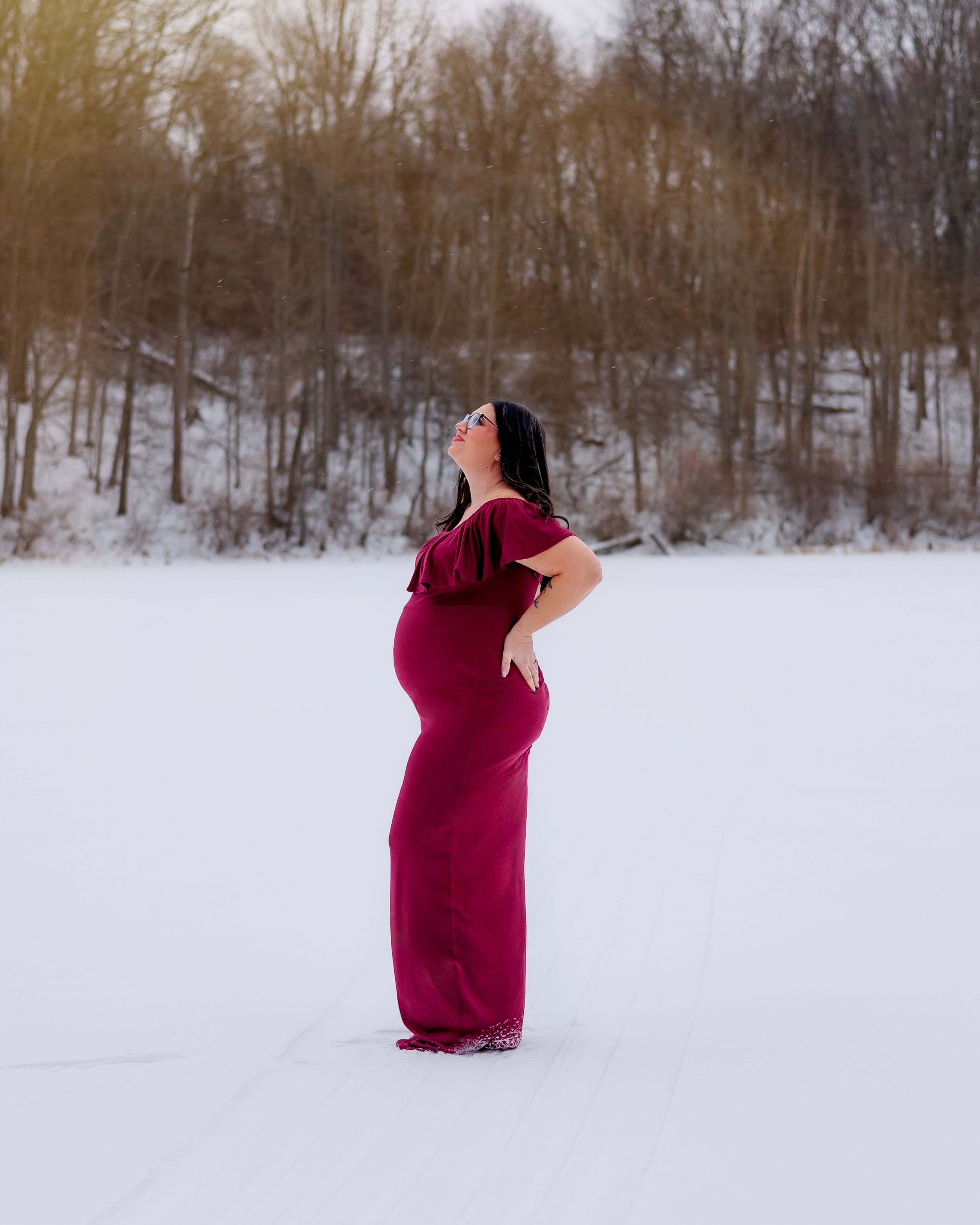 Person in burgundy dress stands in snowy winter landscape with bare trees in background.