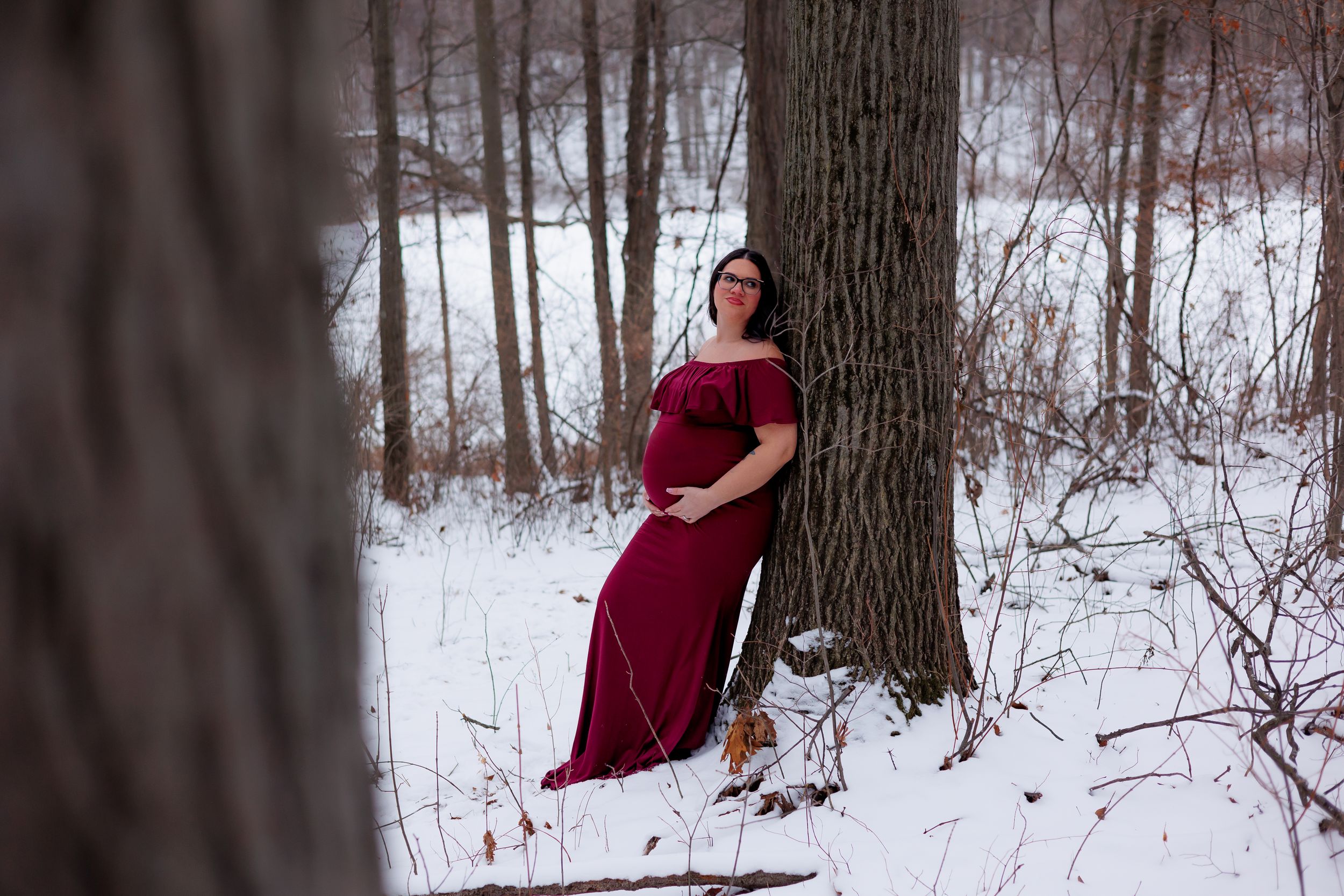 A person in a flowing burgundy dress poses gracefully against snowy winter trees in a forest setting.