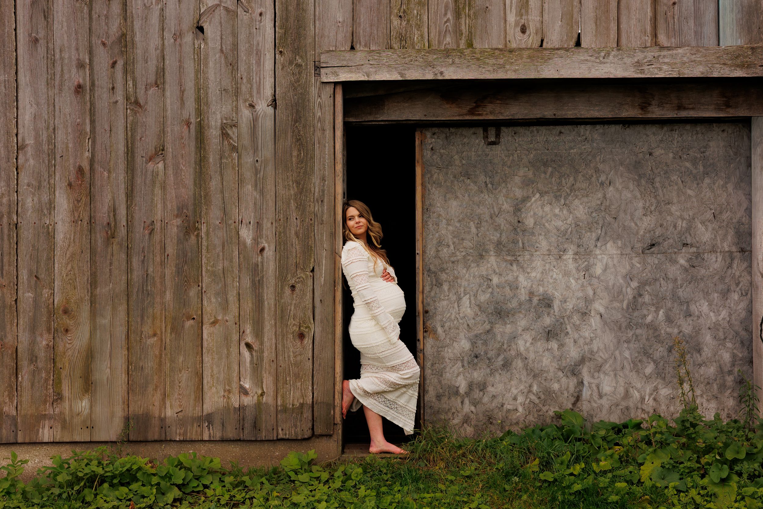 A pregnant woman in a white dress stands in the doorway of a rustic wooden barn.