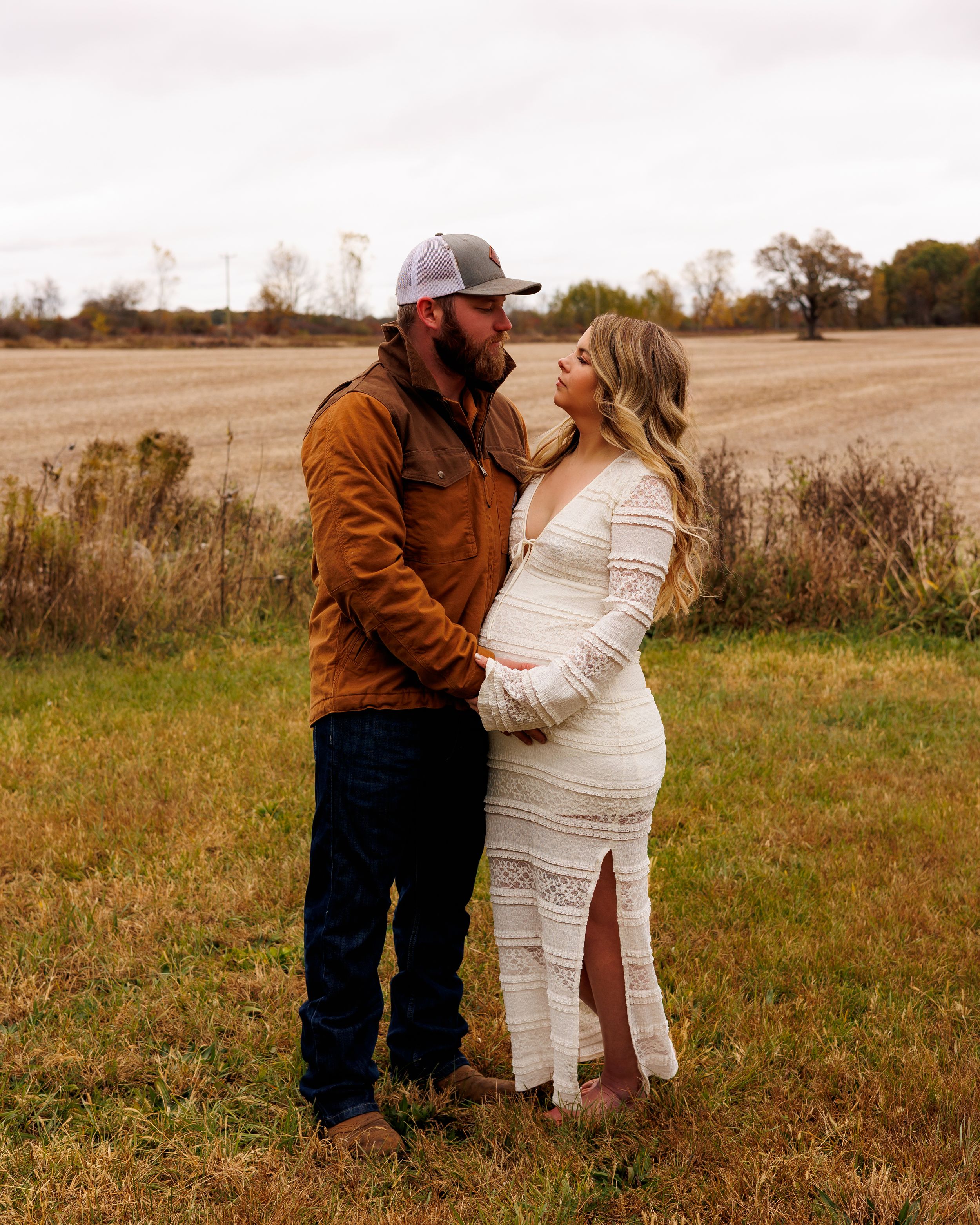 A couple in a field share an intimate moment as she wears a white dress and he wears a brown jacket.