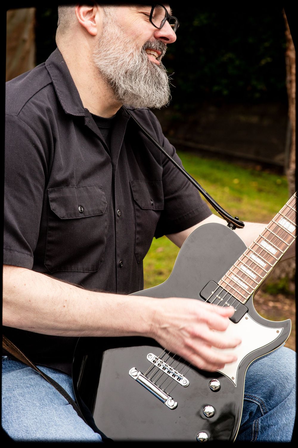 Musician wearing glasses and a black shirt plays a dark electric guitar while smiling outdoors.