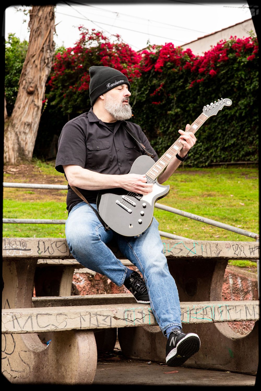 A guitarist in black beanie and shirt plays an electric guitar while sitting on concrete steps near red flowers.
