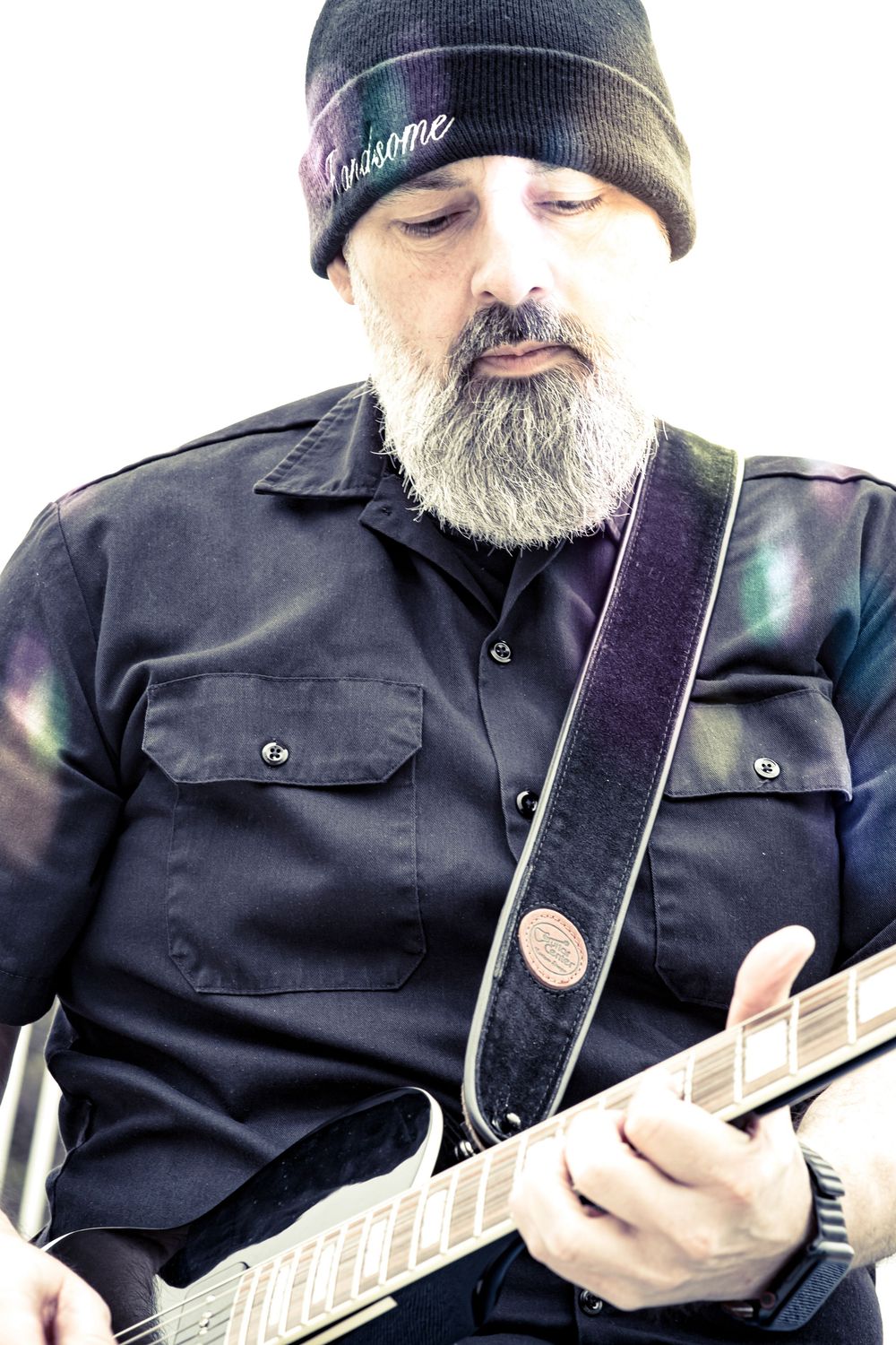 Musician wearing black beanie and button-down shirt plays guitar while looking down at the strings in moody lighting.
