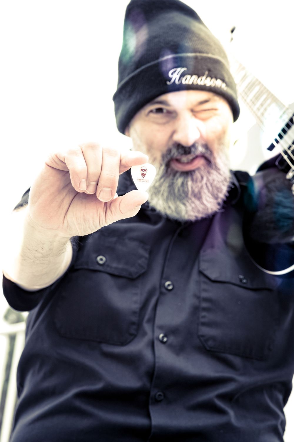 A bearded person in a black beanie hat holds up a small white guitar pick while smiling at the camera.