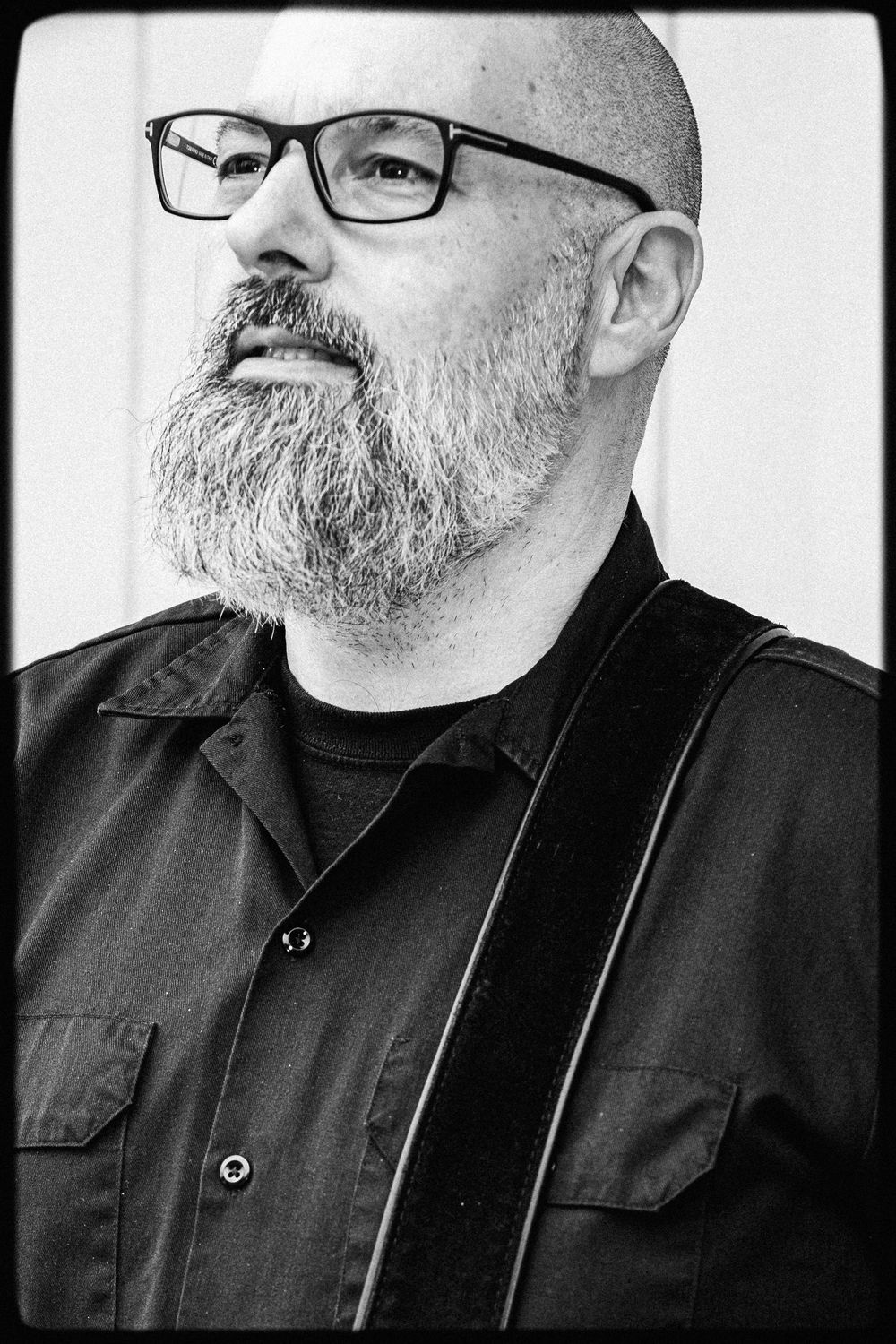 Black and white portrait of a person wearing glasses with a full beard and black buttoned shirt.