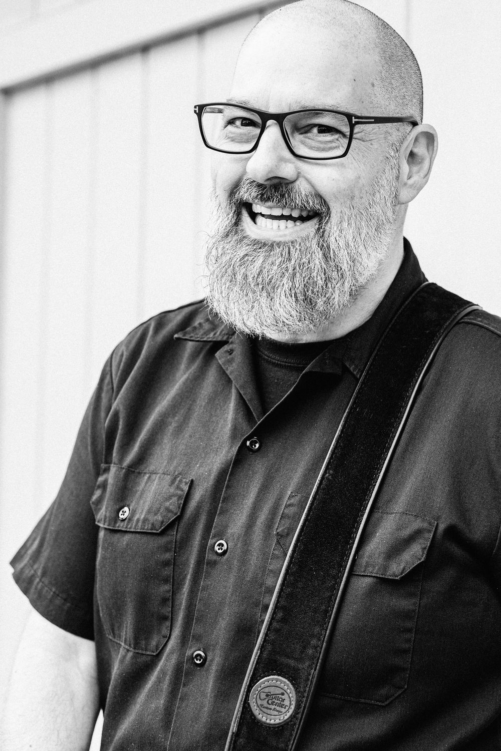Black and white portrait of a bald person with glasses and full beard wearing dark clothing and smiling warmly.