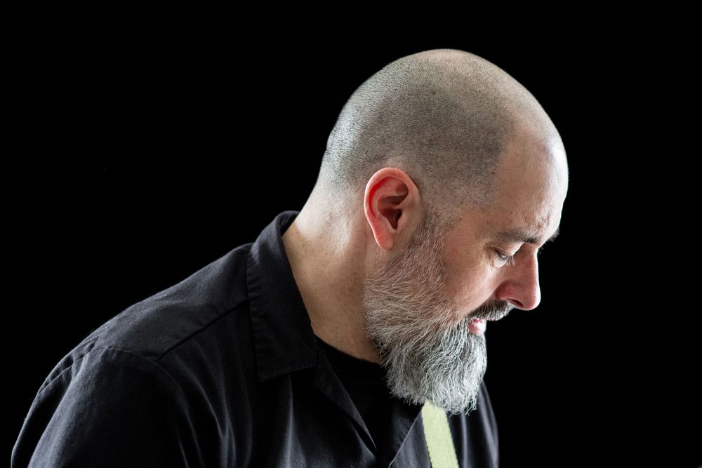 Side profile of a person with a gray beard wearing a black shirt looking downward against a dark background.
