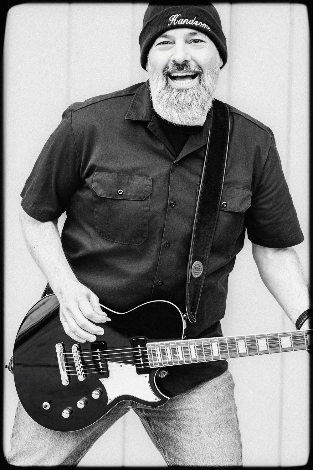 Black and white portrait of a musician with a full white beard wearing a beanie and holding a Les Paul style electric guitar.