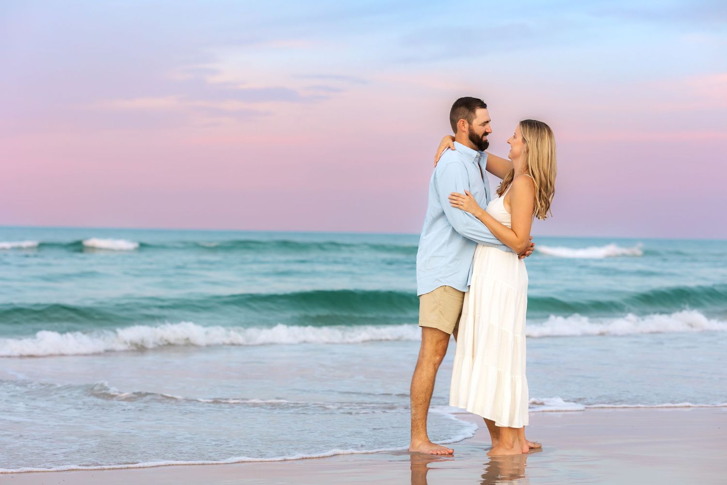 Romantic couple embracing on beach at sunset wearing light summer attire.