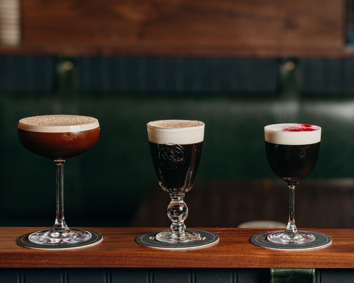 Three elegant cocktail glasses arranged on a dark wooden bar surface with varying colors and garnishes.