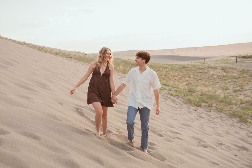 A couple walks hand in hand along scenic sand dunes during sunset at a desert location.