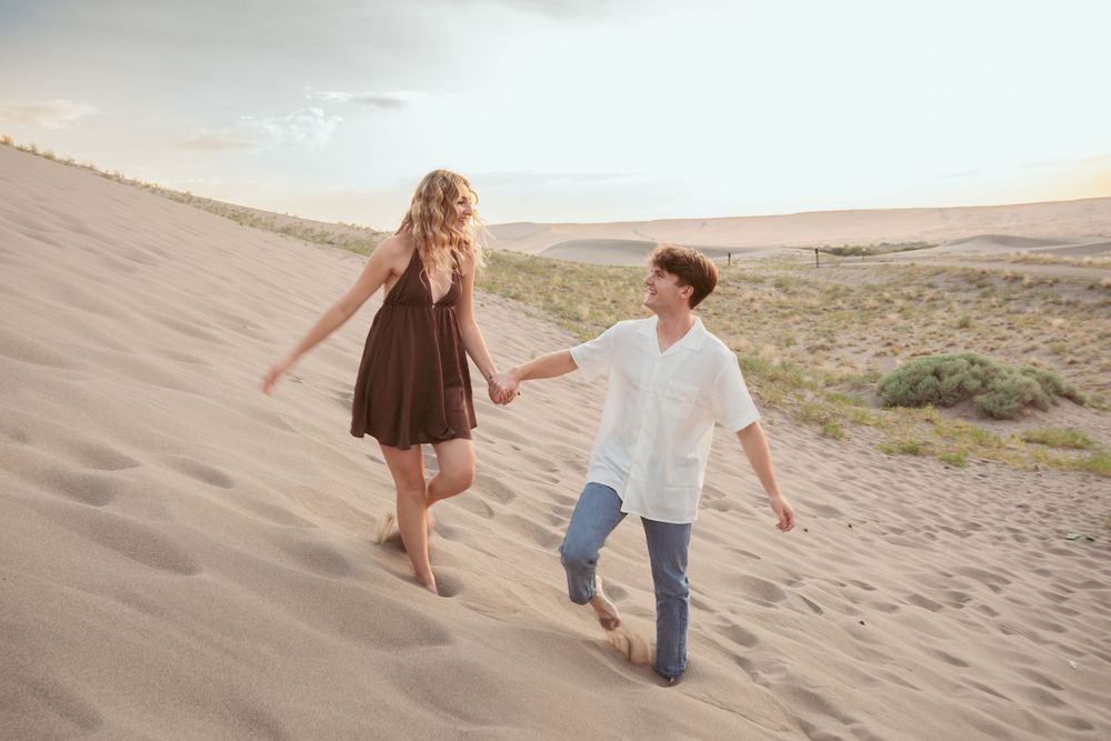 A couple walks hand in hand along sand dunes during golden hour, their silhouettes visible against the setting sun.