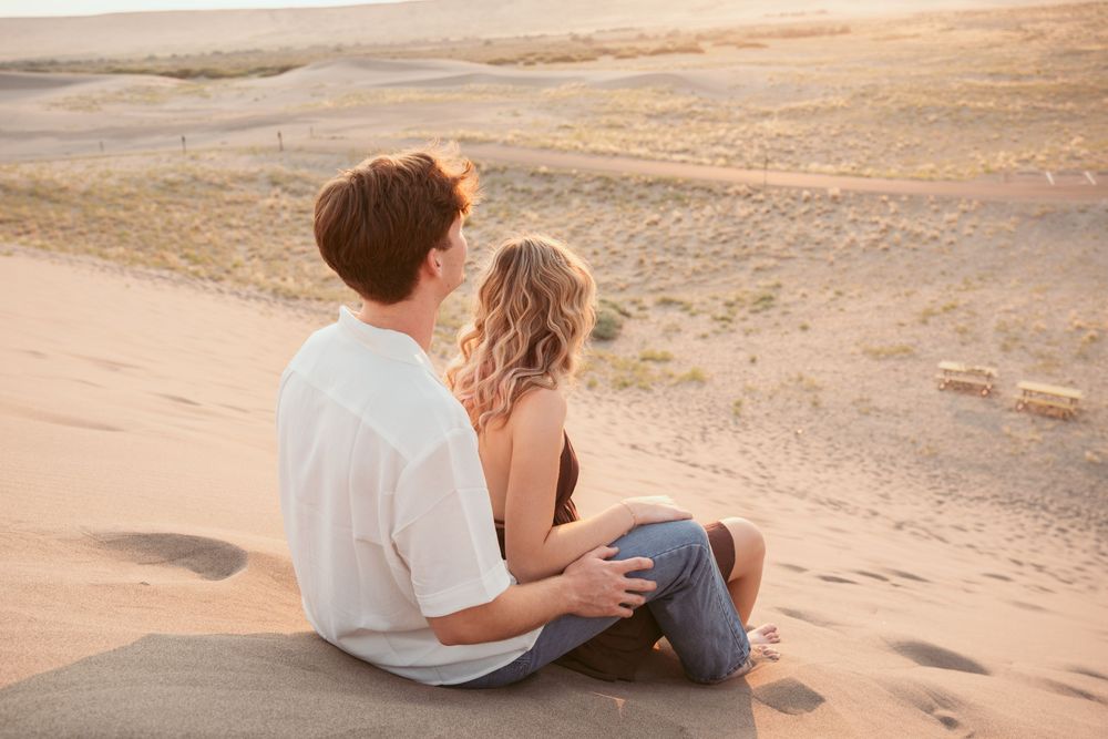 A couple embraces on sandy dunes during golden hour as sunlight casts long shadows across the desert landscape.