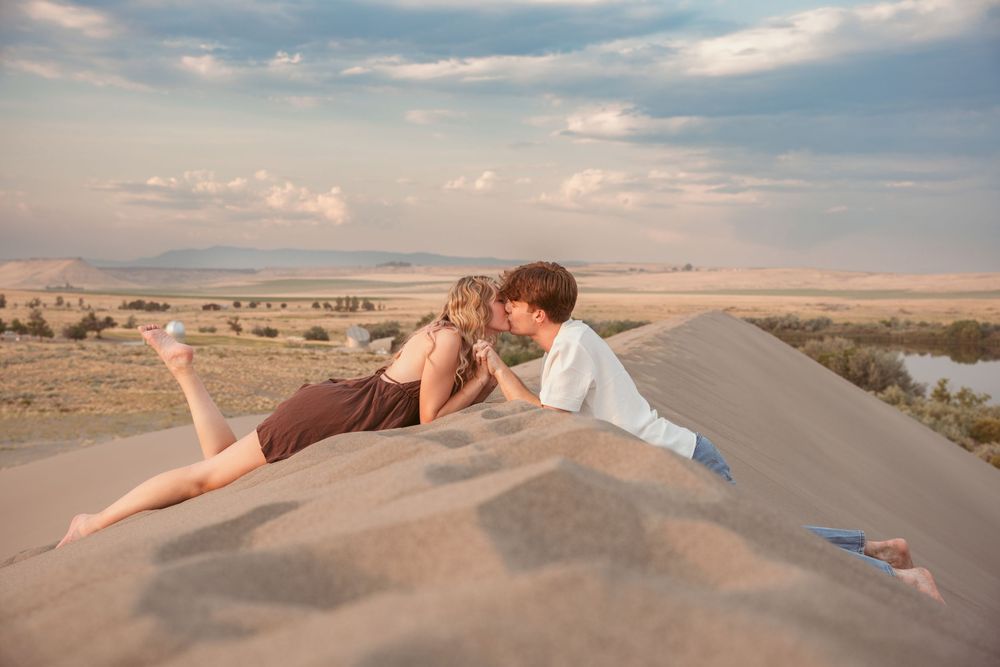Two people relax together on sandy dunes beside an empty road stretching across an arid desert landscape at sunset.