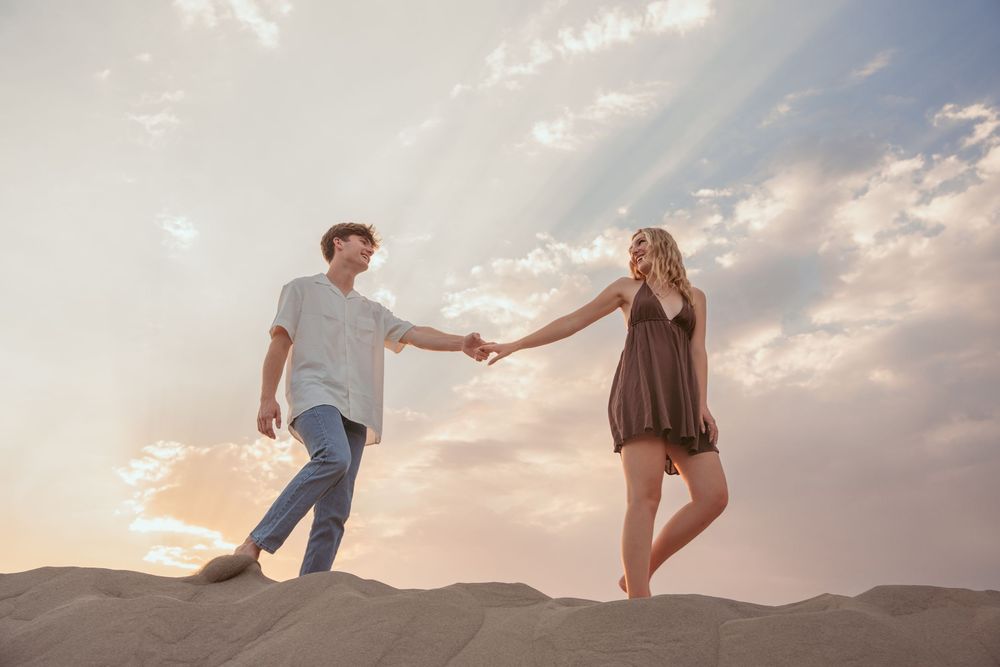 Two people hold hands while standing atop sand dunes at sunset with dramatic clouds in the background.