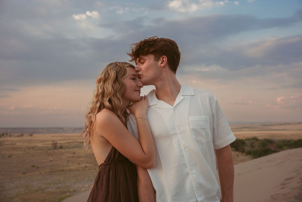 A romantic couple embraces against a desert landscape at sunset with dramatic cloudy skies.