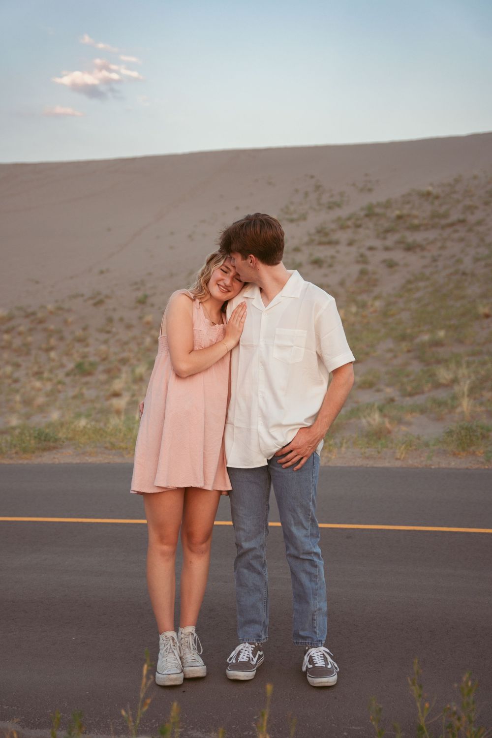 A romantic couple poses together on a desert road at sunset wearing casual clothing and sneakers.