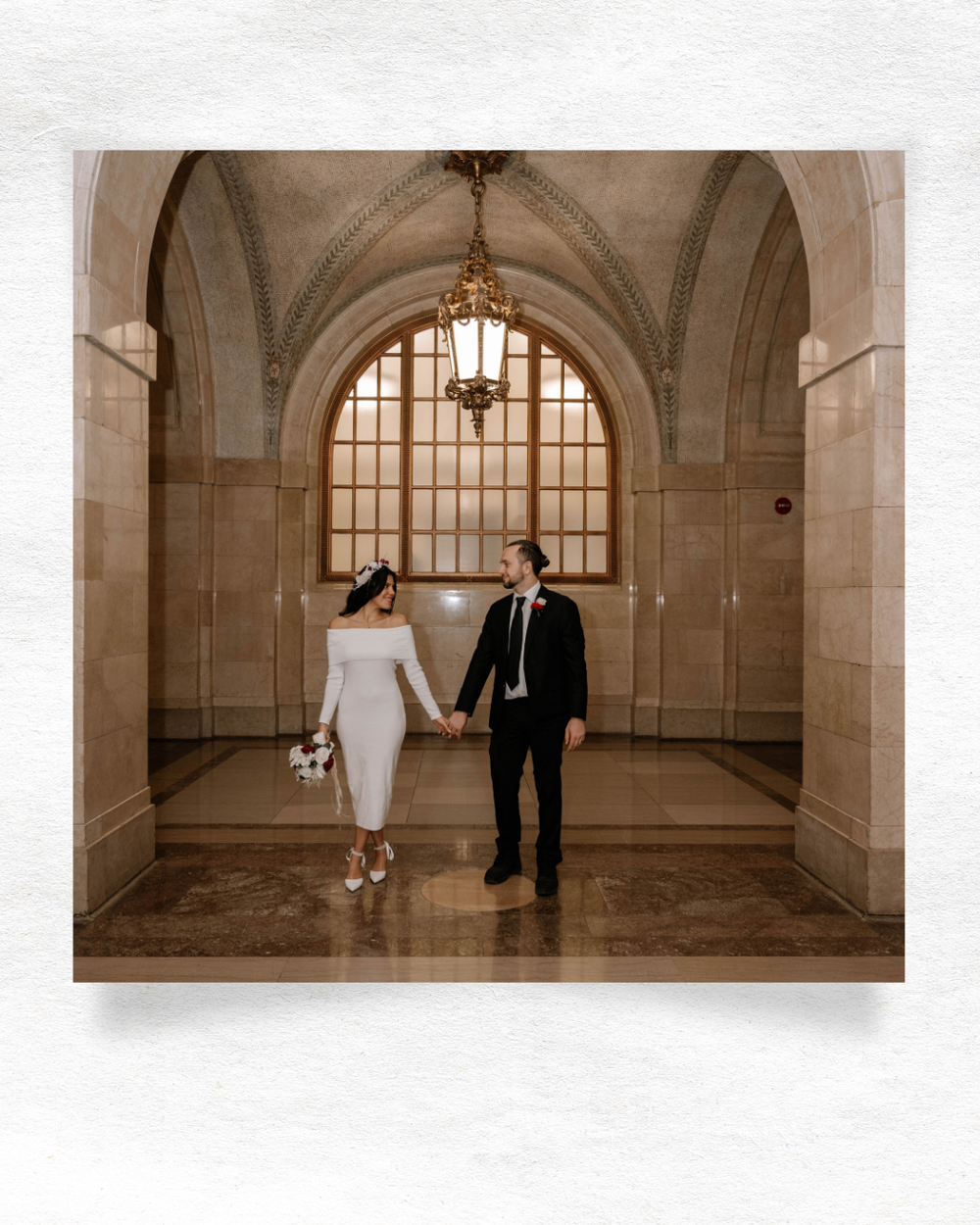 Couple in elegant wedding attire walking hand in hand through an architectural hallway with arched windows and stone walls.