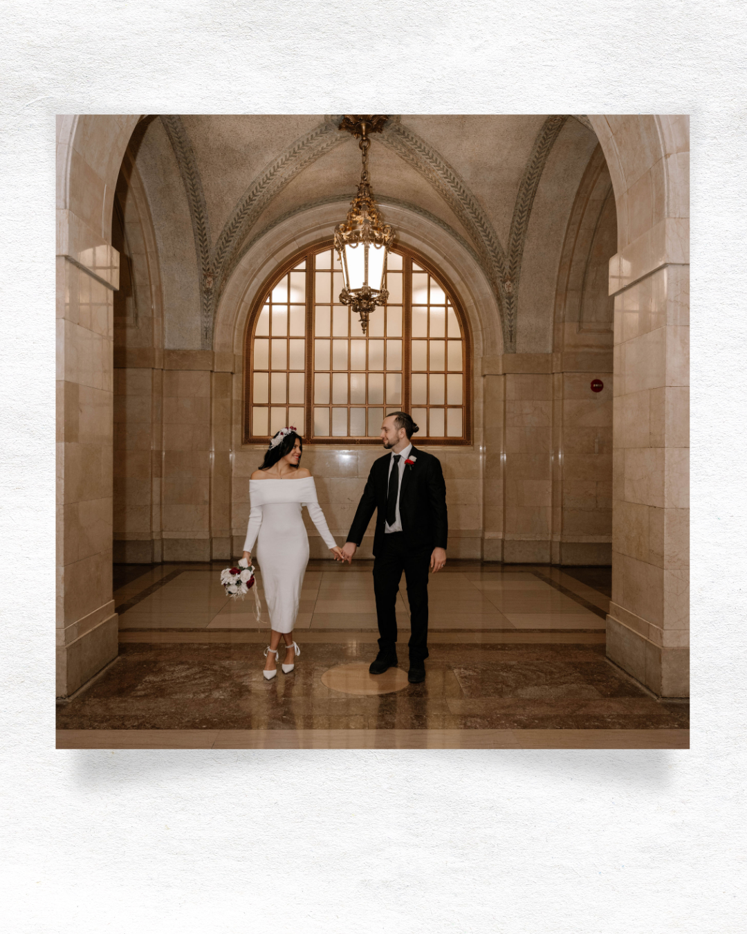 Couple in elegant wedding attire walking hand in hand through an architectural hallway with arched windows and stone walls.