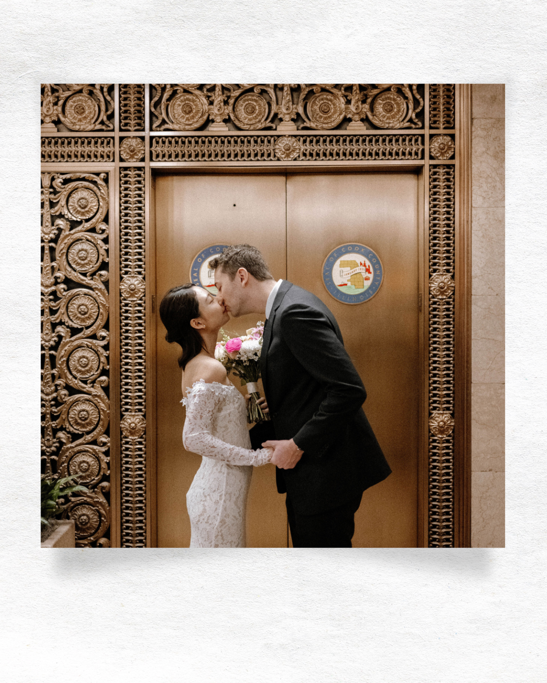 A newlywed couple shares a romantic kiss in front of an ornate elevator door with detailed metalwork.