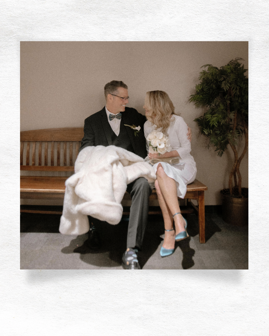A couple sits together on a wooden bench, with one person in a white dress and another in formal attire.