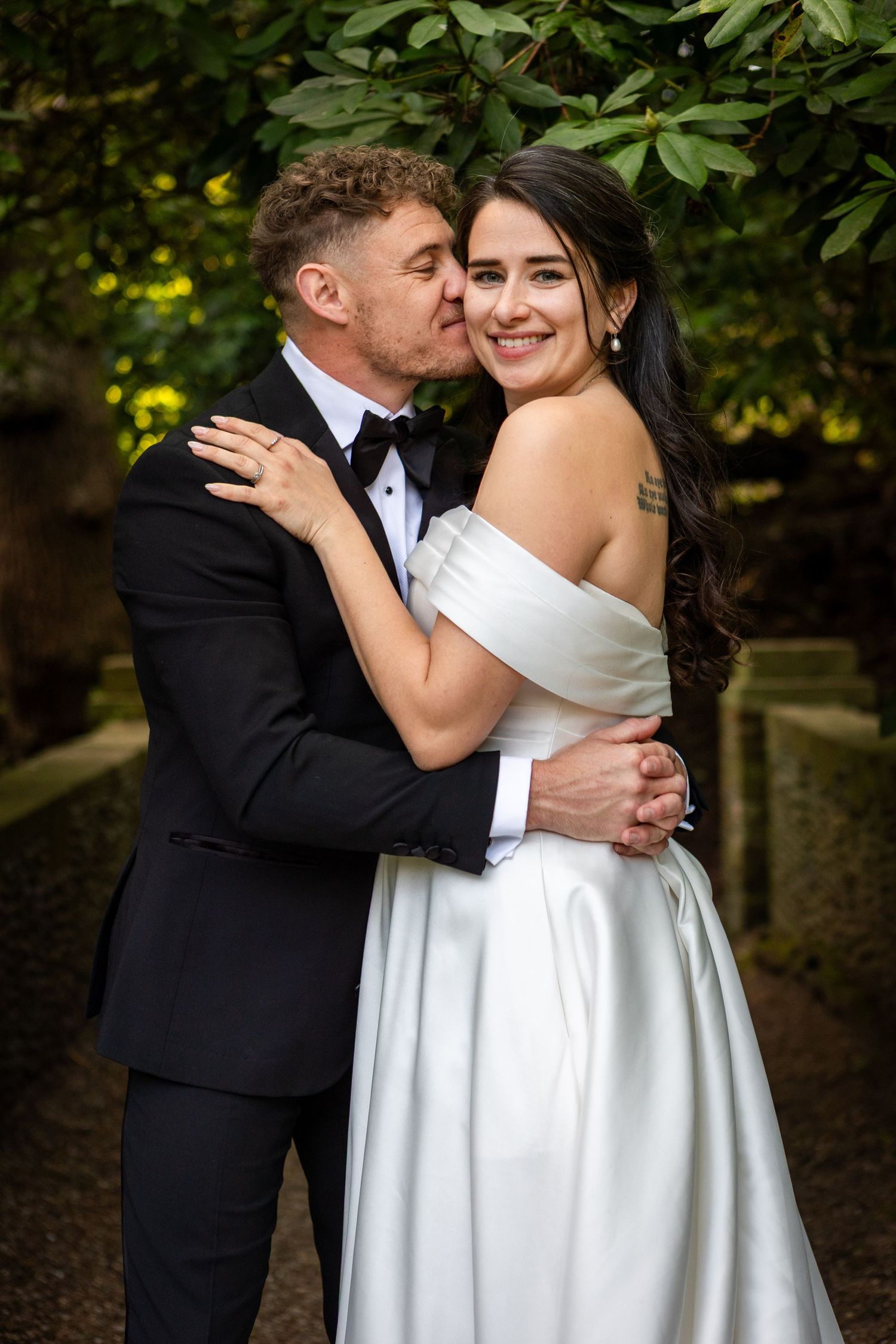 Groom in black tuxedo kisses smiling bride on cheek while embracing her outdoors among lush greenery.