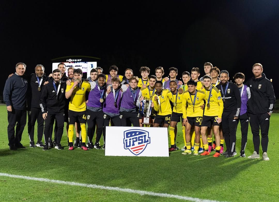 Soccer team in yellow jerseys gathers with coaching staff for group photo with USL league sign on field.
