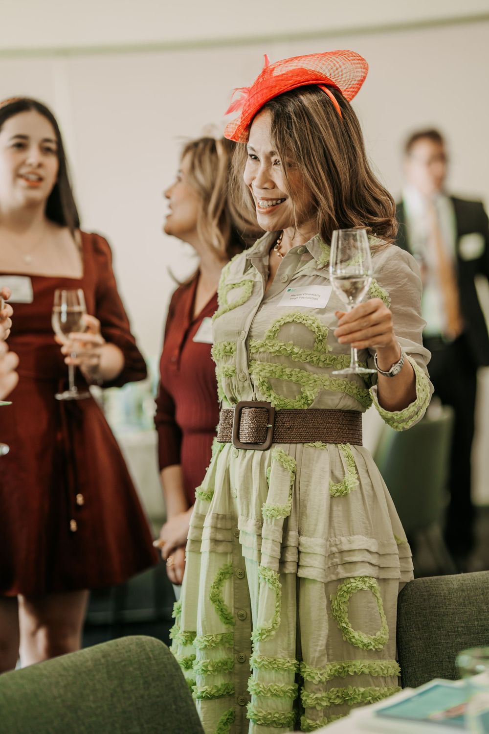 A person in a green vintage dress and orange hat holds champagne at a social gathering.